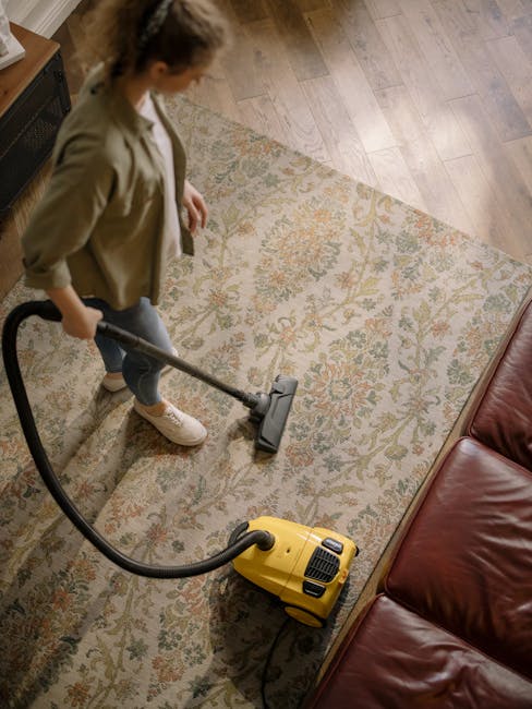 A woman operating a yellow vacuum cleaner with a black hose on a patterned area rug in a living room. The rug features a floral design in muted tones of beige, green, and orange, placed over a wooden floor. The woman is wearing a beige jacket, white shoes, and standing next to a red leather sofa. Natural light illuminates the space, highlighting the cleanliness of the rug and the wooden flooring. The scene depicts domestic surface cleaning, emphasizing the professional services of Holland Park Carpet Cleaners for maintaining hygiene and surface hygiene in residential settings.